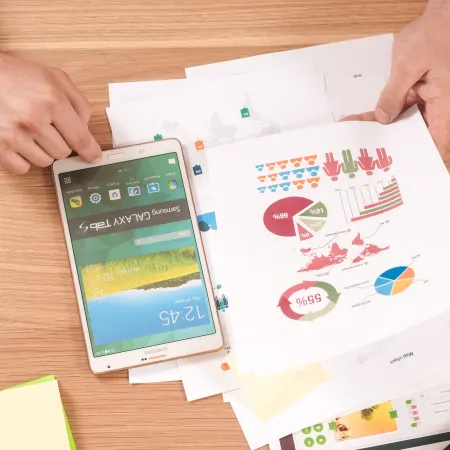 Hands holding a Samsung Galaxy Tab and colorful business charts on a wooden desk with office supplies.