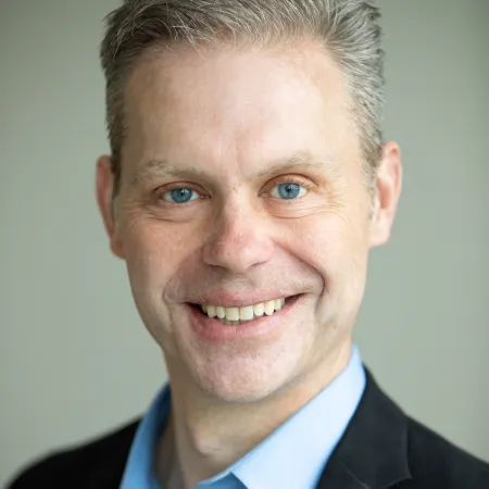 Smiling middle-aged man with blue eyes wearing a light blue shirt and black blazer.