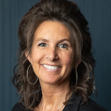 Smiling woman with curly hair wearing hoop earrings, a necklace, a black blazer, and a maroon top against dark background
