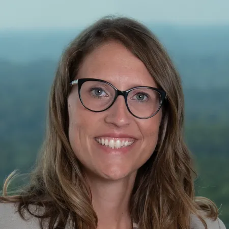Smiling woman with glasses and wavy brown hair wearing a beige blazer, posed against a blurred natural background