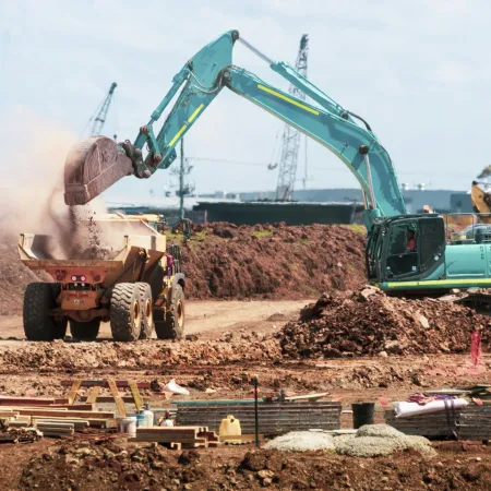 Excavator loading dirt into dump truck at active construction site with cranes and materials around.