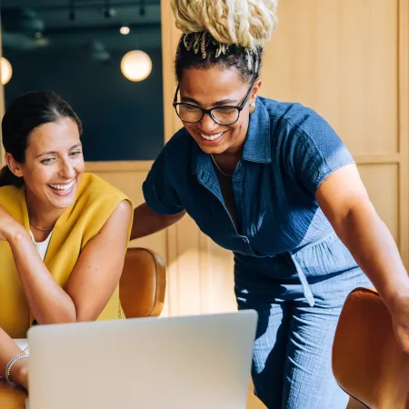 Two women smiling and collaborating while looking at a laptop in a modern office setting