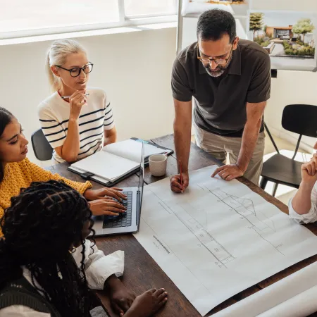 Architect team collaborating over building blueprints in a bright office with laptops and coffee cups.