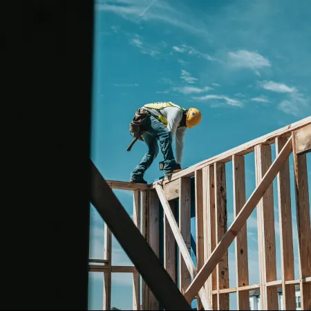 Construction worker in a yellow helmet working on wooden framing against a clear blue sky.