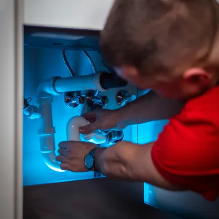 Plumber in red shirt fixing under-sink white PVC pipes illuminated by blue light inside kitchen cabinet.