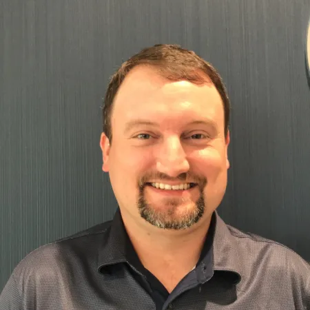 Smiling man with short brown hair and goatee wearing a dark gray polo shirt against a textured dark background