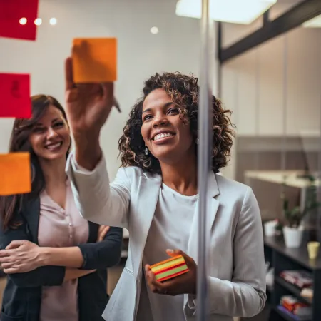 Two businesswomen smiling and placing sticky notes on a glass wall during a brainstorming session in an office.