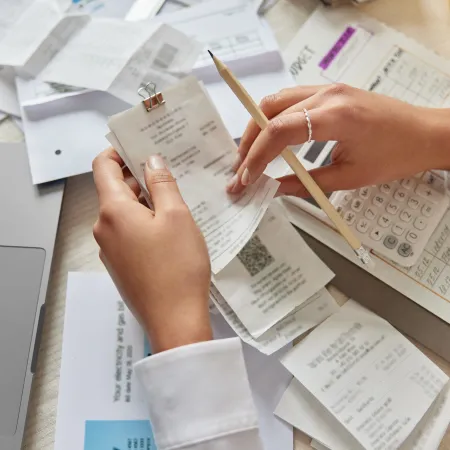 Person organizing receipts with pencil and calculator on a cluttered desk with laptop and documents.