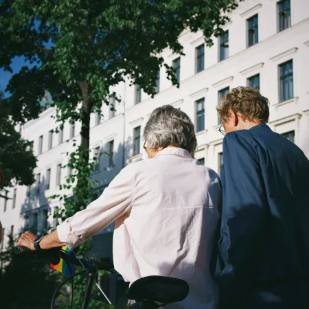 Two people standing next to a bicycle on a sunny street with white buildings and green trees.
