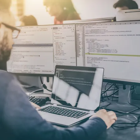 Software developer working on code displayed on laptop and dual monitors in modern office.