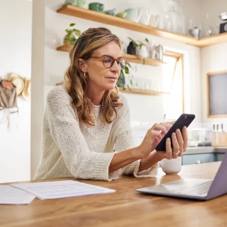 Woman in glasses using smartphone while working on laptop at wooden kitchen table with documents and mug.