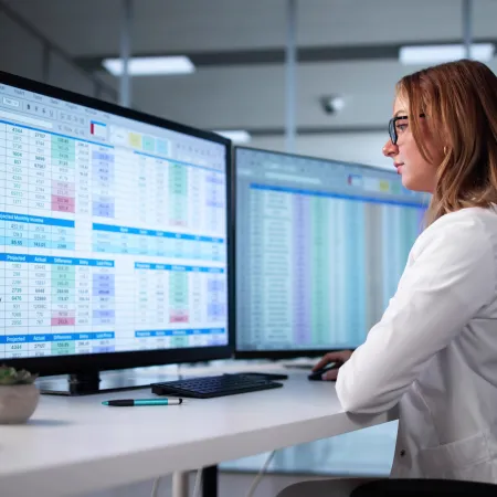 Professional woman analyzing data on dual monitors in modern office environment with spreadsheets and charts