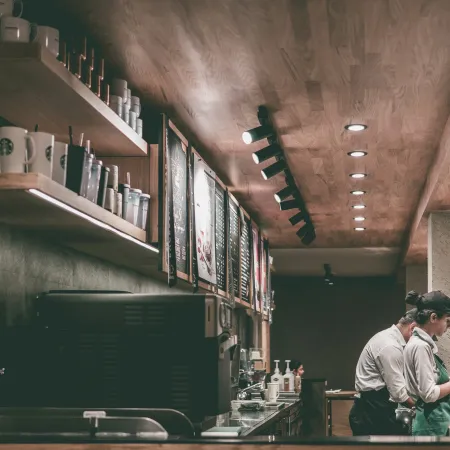 Baristas preparing drinks behind counter in cozy coffee shop with wooden ceiling and shelves of mugs