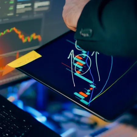Person pointing at digital tablet displaying candlestick charts and financial graphs on computer screens in background