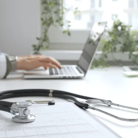 Stethoscope on medical chart in foreground with doctor typing on laptop in bright office setting.