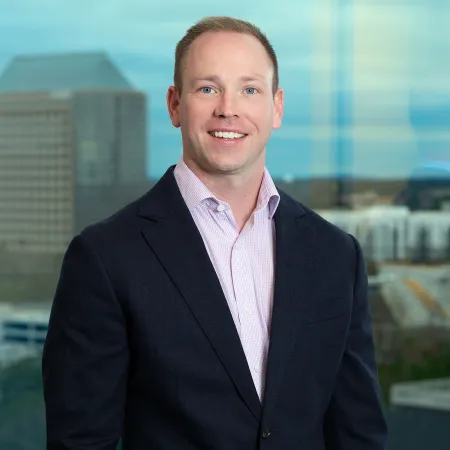 Smiling man in business attire with a cityscape background, wearing a dark blazer and light pink shirt.