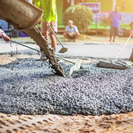 Workers pouring and leveling wet concrete on ground during road construction under sunlight.