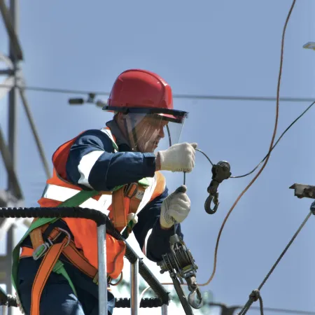Electrician in safety gear working on power lines with a red hard hat and orange harness on a clear day.