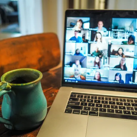 Laptop showing a video conference with multiple participants next to a green ceramic coffee mug on a wooden table.