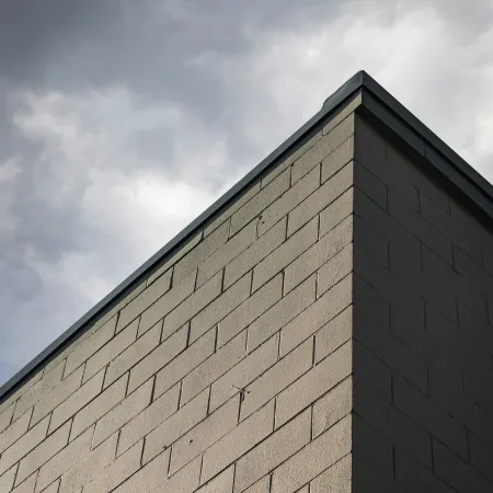 Corner of a beige brick building with a dark metal roof edge under a cloudy sky.
