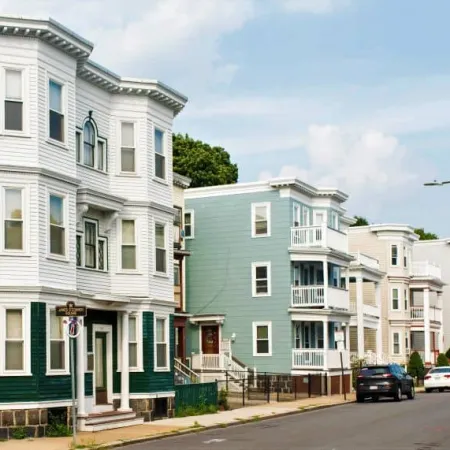 Row of multi-story residential houses with balconies along a quiet street under a blue sky.