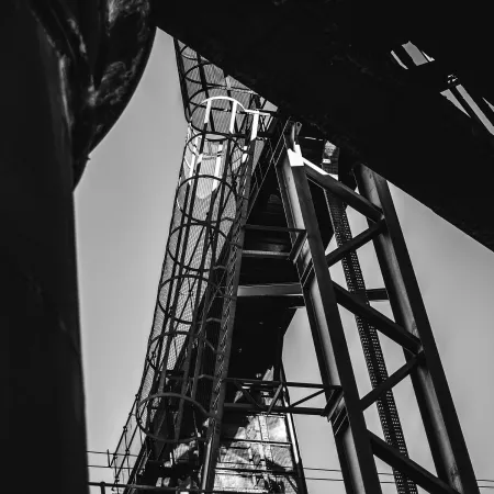 Black and white upward view of industrial metal framework and staircases with safety cages against the sky.