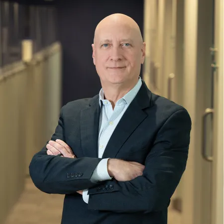 Confident bald man in navy suit with arms crossed standing in modern office hallway with glass doors.