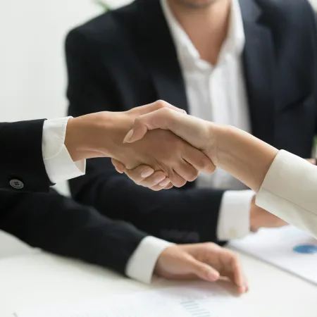 Two business professionals shaking hands across a white table during a meeting, showing cooperation and agreement.
