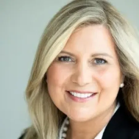 Portrait of a smiling woman with blonde hair, wearing a black blazer and pearl necklace against neutral background