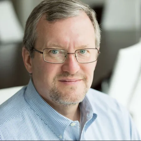 Middle-aged man with glasses and beard wearing a blue striped shirt sitting in a bright office room.