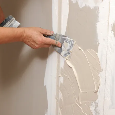 Hands applying plaster on drywall with putty knives during wall repair or finishing work.