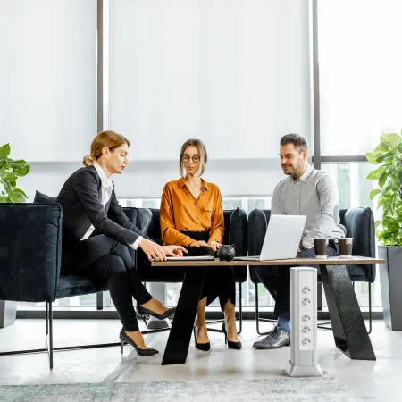 Three professionals collaborate around a laptop in a modern office with large windows and plants.