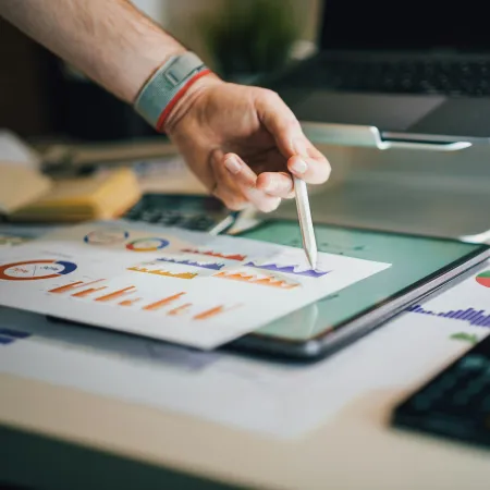Person analyzing colorful charts on paper and tablet with a laptop and calculator on a desk
