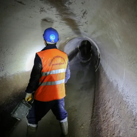 Worker in orange vest and blue helmet inspecting a dimly lit tunnel with reflective water on the floor.