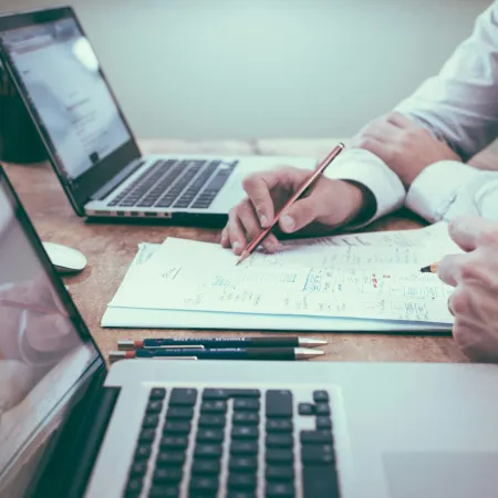 Two people working with laptops and documents on a wooden table, discussing and taking notes.