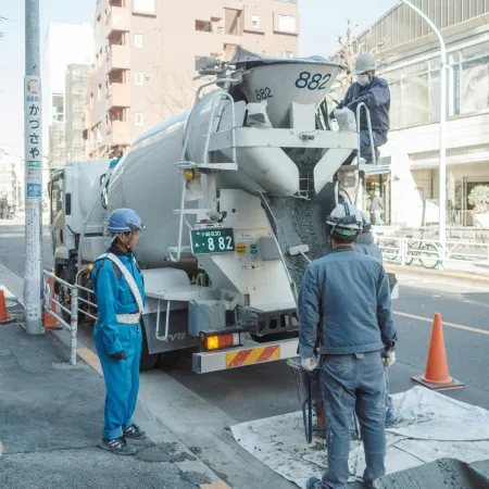 Construction workers operating a concrete mixer truck on a city street near orange safety cones and buildings.
