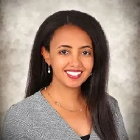 Smiling woman with long dark hair wearing gray top and pearl earrings against a blurred gray background