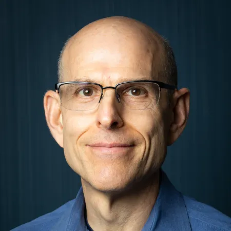 Middle-aged man with glasses and a blue shirt smiling against a dark blue background.
