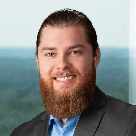Smiling man with red beard wearing a blue shirt and dark suit jacket in front of a blurred natural background