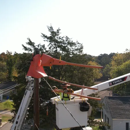 Utility worker in bucket truck repairing power lines on wooden pole in suburban neighborhood with autumn trees.