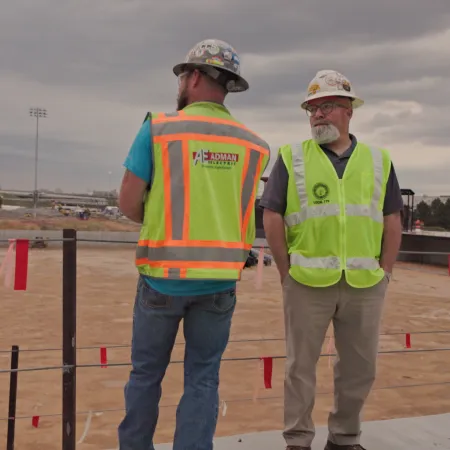 Two construction workers wearing hard hats and safety vests at a large outdoor construction site under cloudy sky.