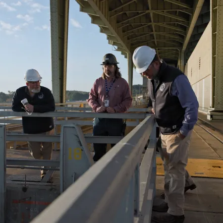 Engineers wearing hard hats inspect a large industrial facility under a bridge on a sunny day.