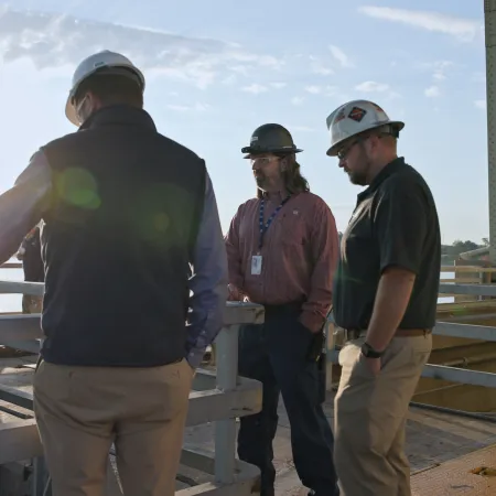Three construction workers wearing helmets discuss plans on a bridge structure during sunset with a lake in the background