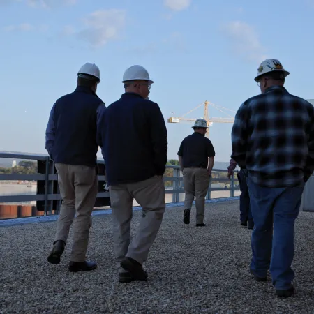 Four construction workers wearing hard hats walking on a gravel rooftop near industrial buildings in daylight