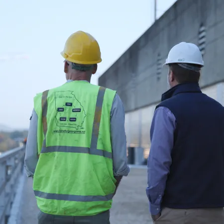 Two construction workers wearing safety helmets and vests walk along an industrial site near a concrete wall.
