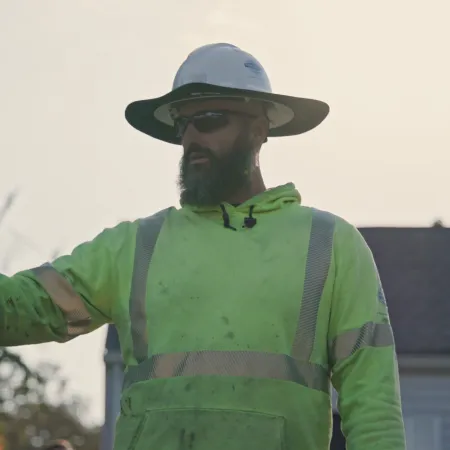 Construction worker wearing a safety helmet and neon green reflective jacket giving instructions outdoors near a house.