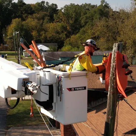Utility worker in bucket lift repairing electrical lines on wooden pole with trees and buildings in background