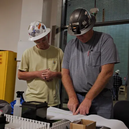 Two construction workers in hard hats reviewing blueprints and plans inside an industrial work area.