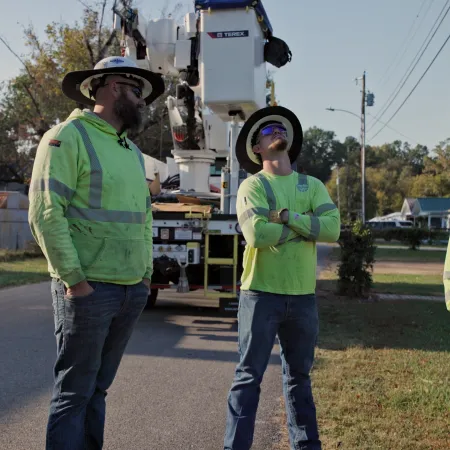 Three utility workers in safety gear standing near a bucket truck on a residential street during daytime.