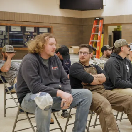 Group of young men in casual workwear seated in a room with ladders and framed certificates on the wall.